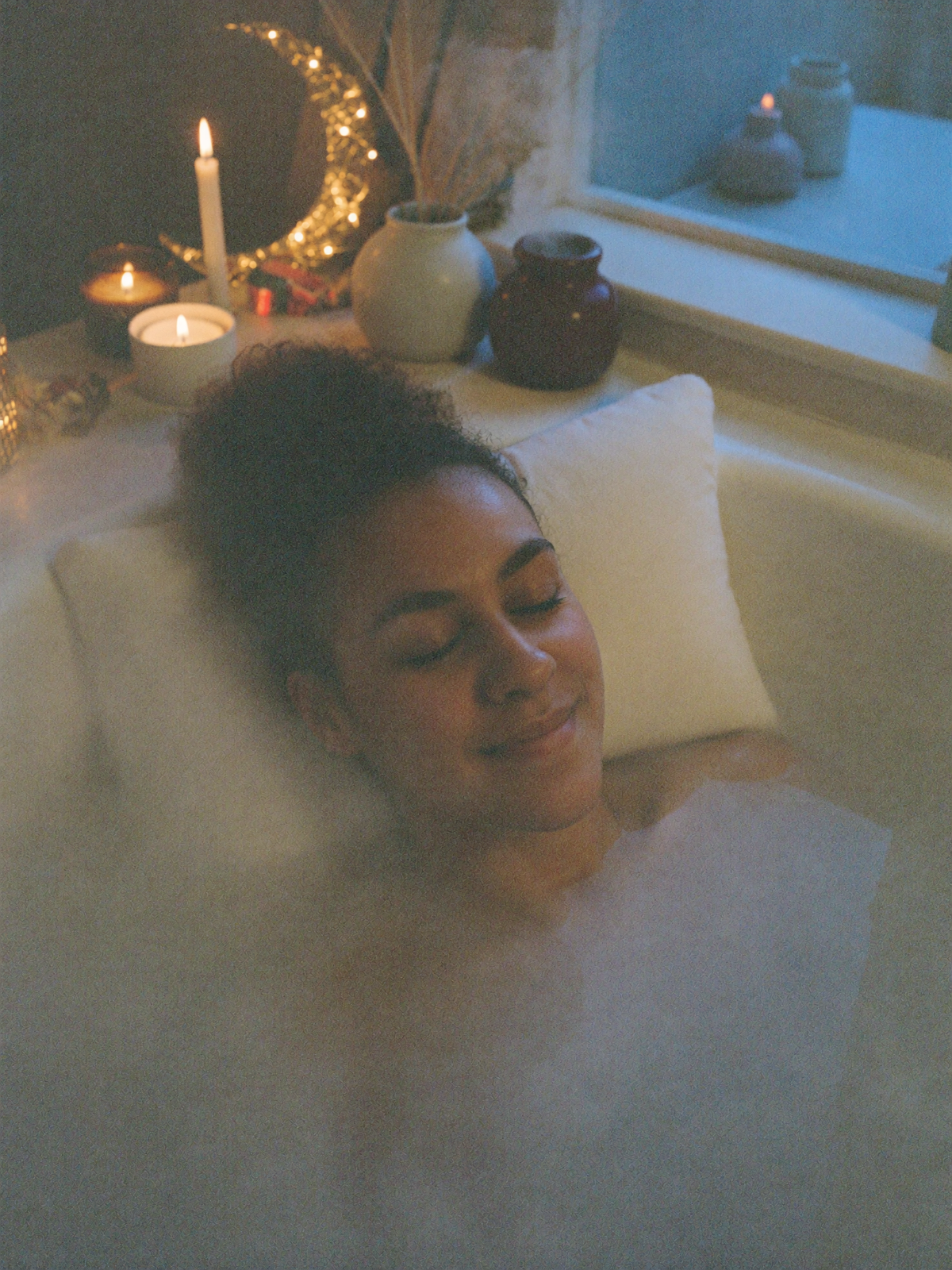 Lisa détendue dans un bain laiteux le soir, la tête sur un coussin de bain, bougies et lune dorée